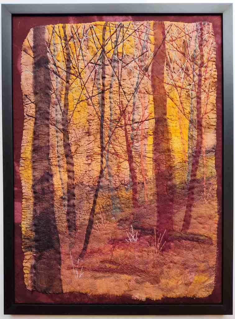 Framed textile artwork with golden sun streaming through trees from behind.